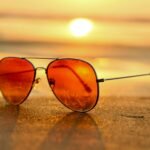 Close-up of sunglasses on a beach during a vibrant sunset, capturing the essence of summer.